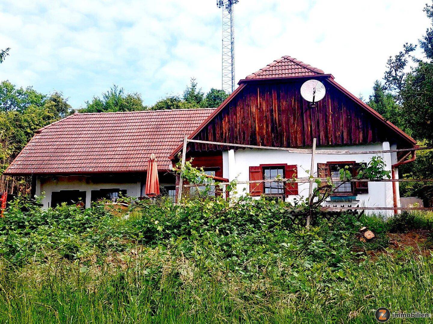 Traditionelles Haus mit rotem Ziegeldach und Holzgiebel, umgeben von üppigem Grün in ländlicher Umgebung.