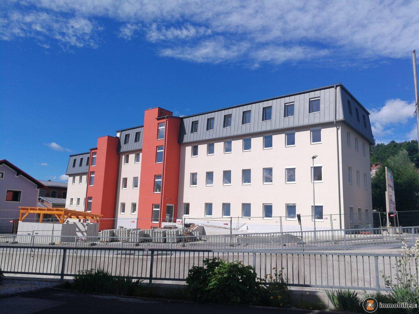 Modernes Mehrfamilienhaus mit beiger und roter Fassade sowie Mansarddach, umgeben von einer Baustelle.