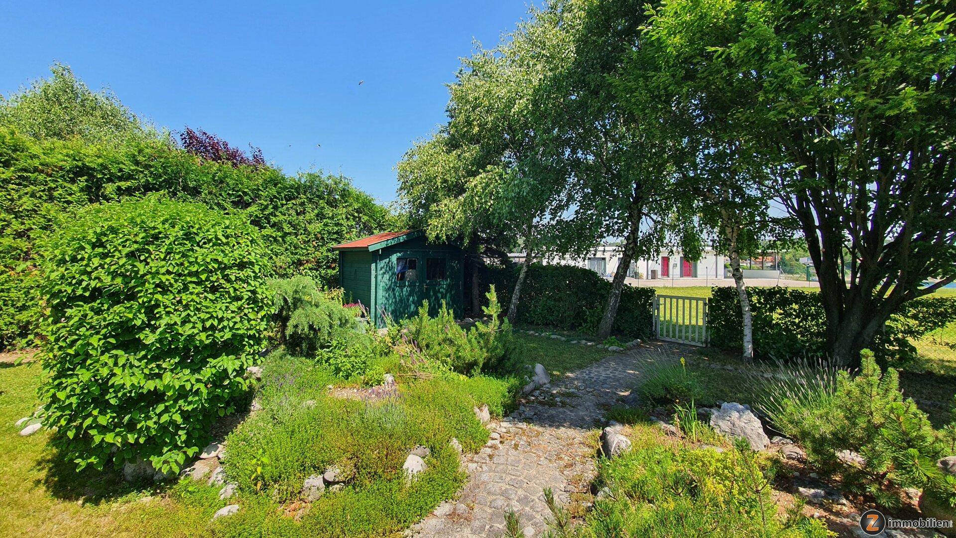 Gartenbereich mit grünem Gartenhaus, hohen Bäumen und einer dichten Hecke, die das Grundstück begrenzt.
