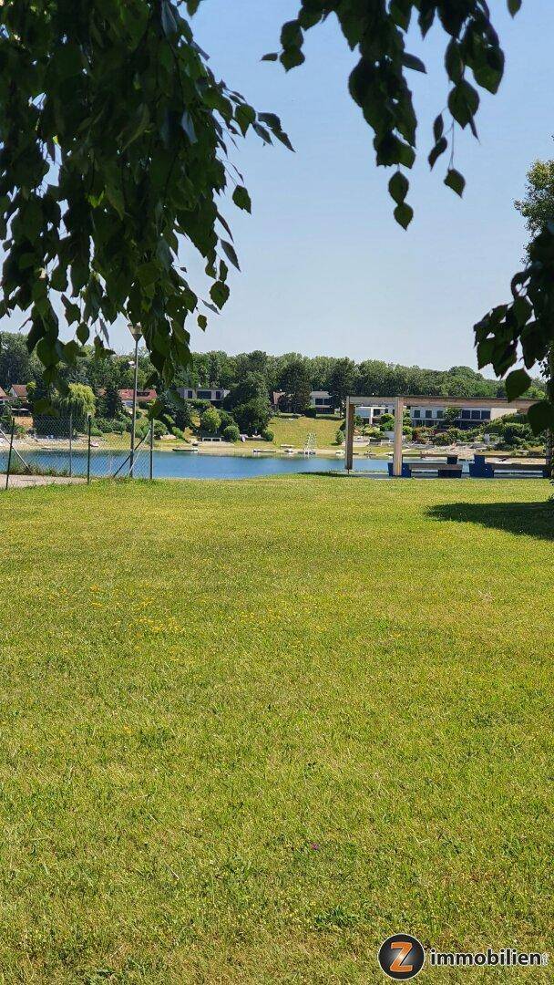 Weitläufige Rasenfläche mit Blick auf einen See und die gegenüberliegende Uferbebauung unter blauem Himmel.