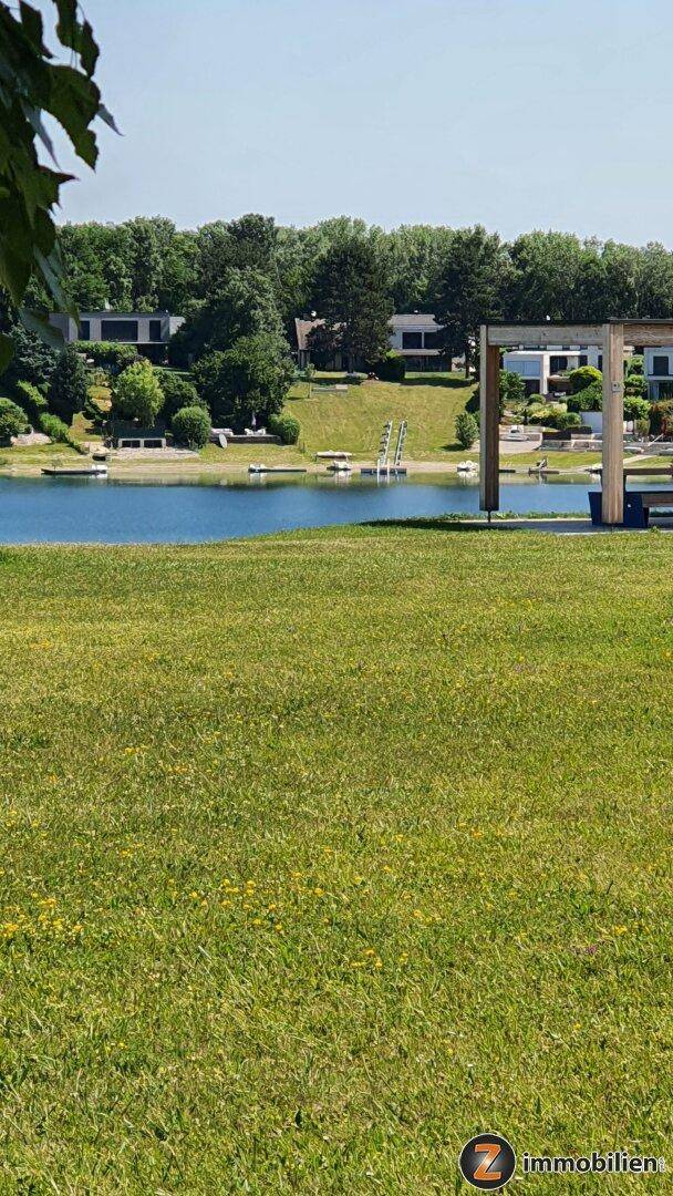 Grüne Rasenfläche mit weitem Blick auf den See und die modernen Häuser am gegenüberliegenden Ufer.