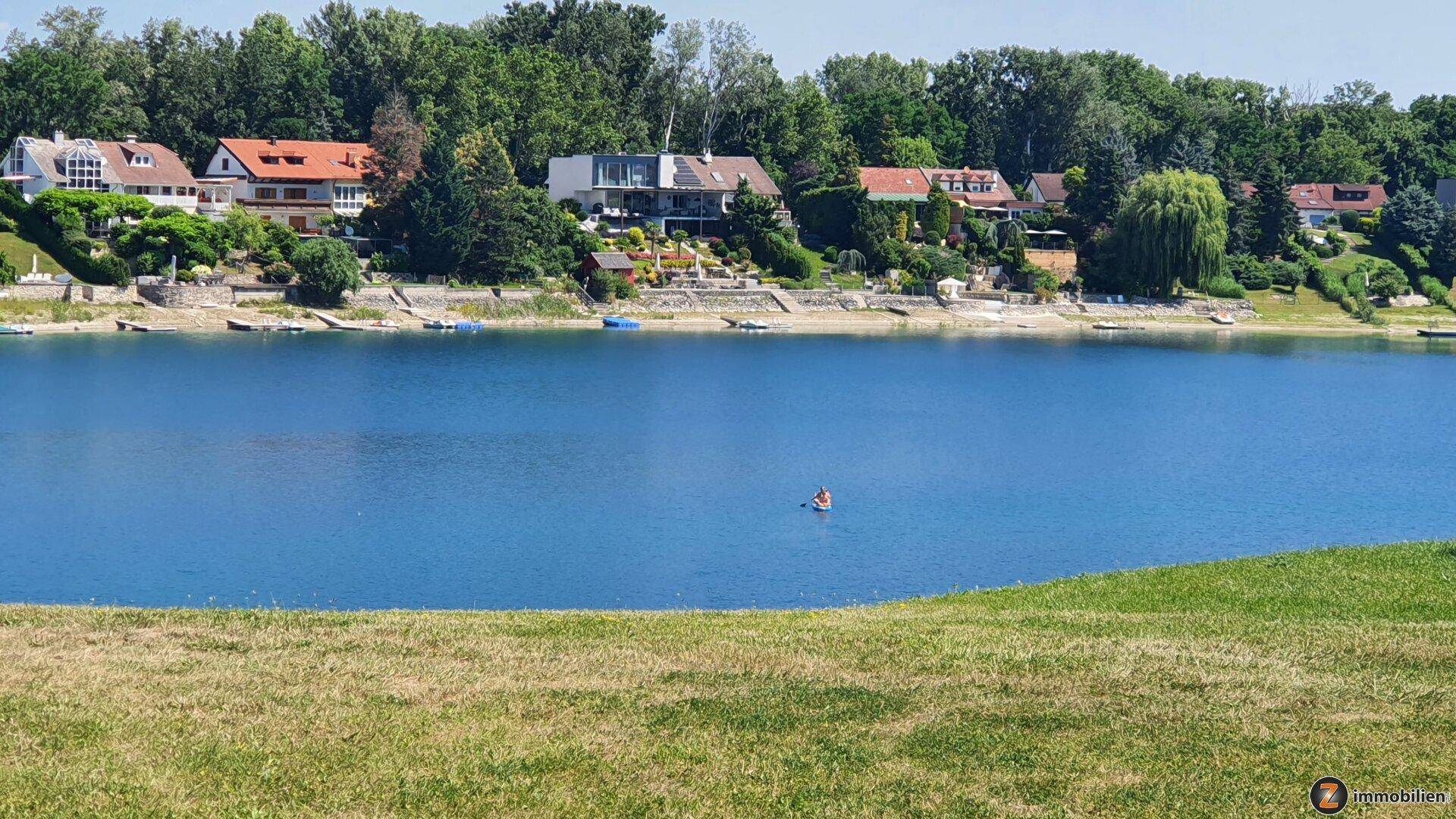 Panoramablick auf einen blauen See mit Häusern am Ufer und einer Person im Wasser, umgeben von Grün.