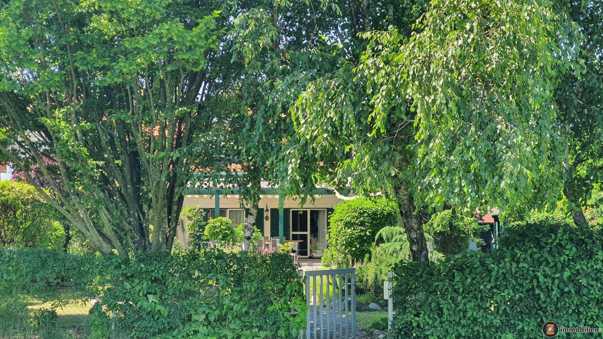 Vorderansicht eines Hauses, umgeben von üppigem Grün, Bäumen und einer Hecke mit einem Gartentor.