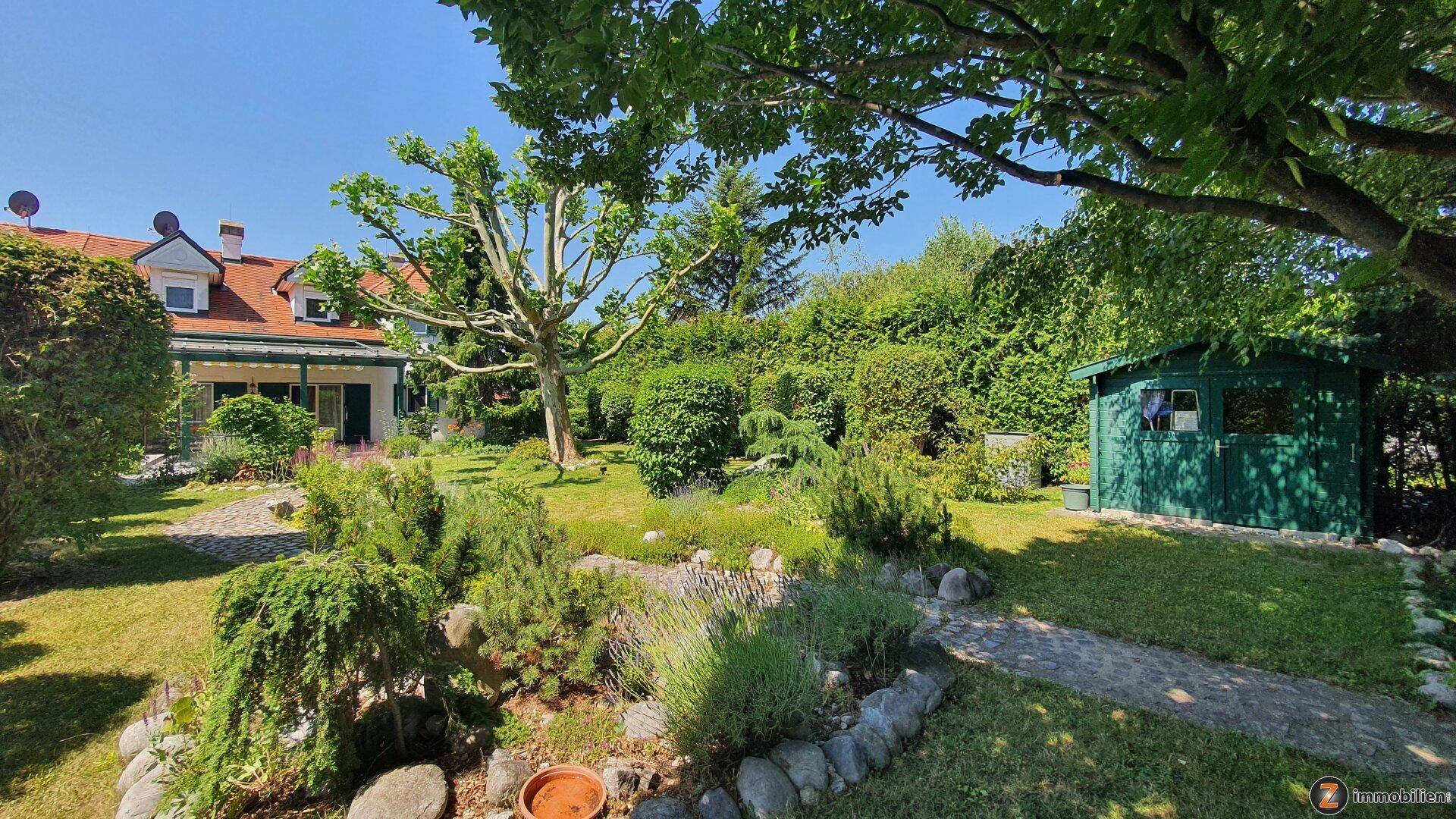 Gepflegter Garten mit Rasenfläche, vielfältiger Bepflanzung, Steinweg und einem kleinen grünen Gartenhaus.