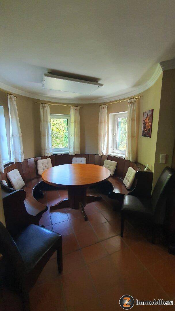 Gemütlicher Essbereich mit rundem Holztisch und eingebauter Sitzbank, umgeben von Fenstern und Terrakottafliesen.