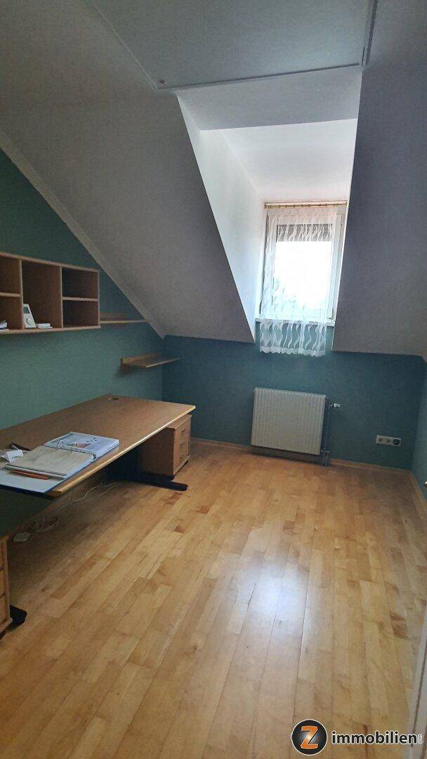 Dachzimmer als Büro genutzt, mit Dachschräge, Schreibtisch, Regalen und einem Fenster für natürliches Licht.