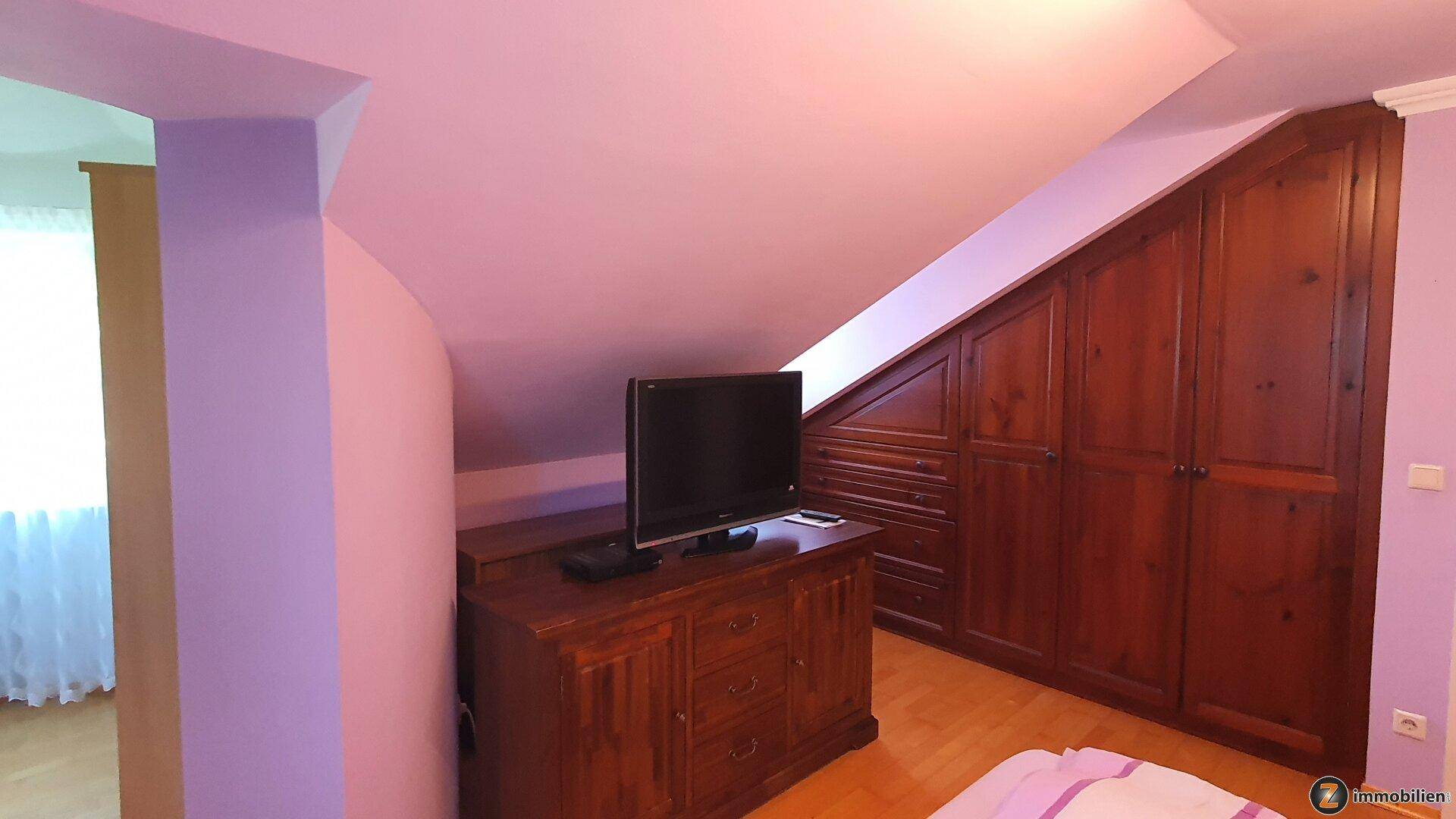 Schlafzimmer mit Fernseher auf einer Holzkommode und maßgefertigten Einbauschränken unter der Dachschräge.