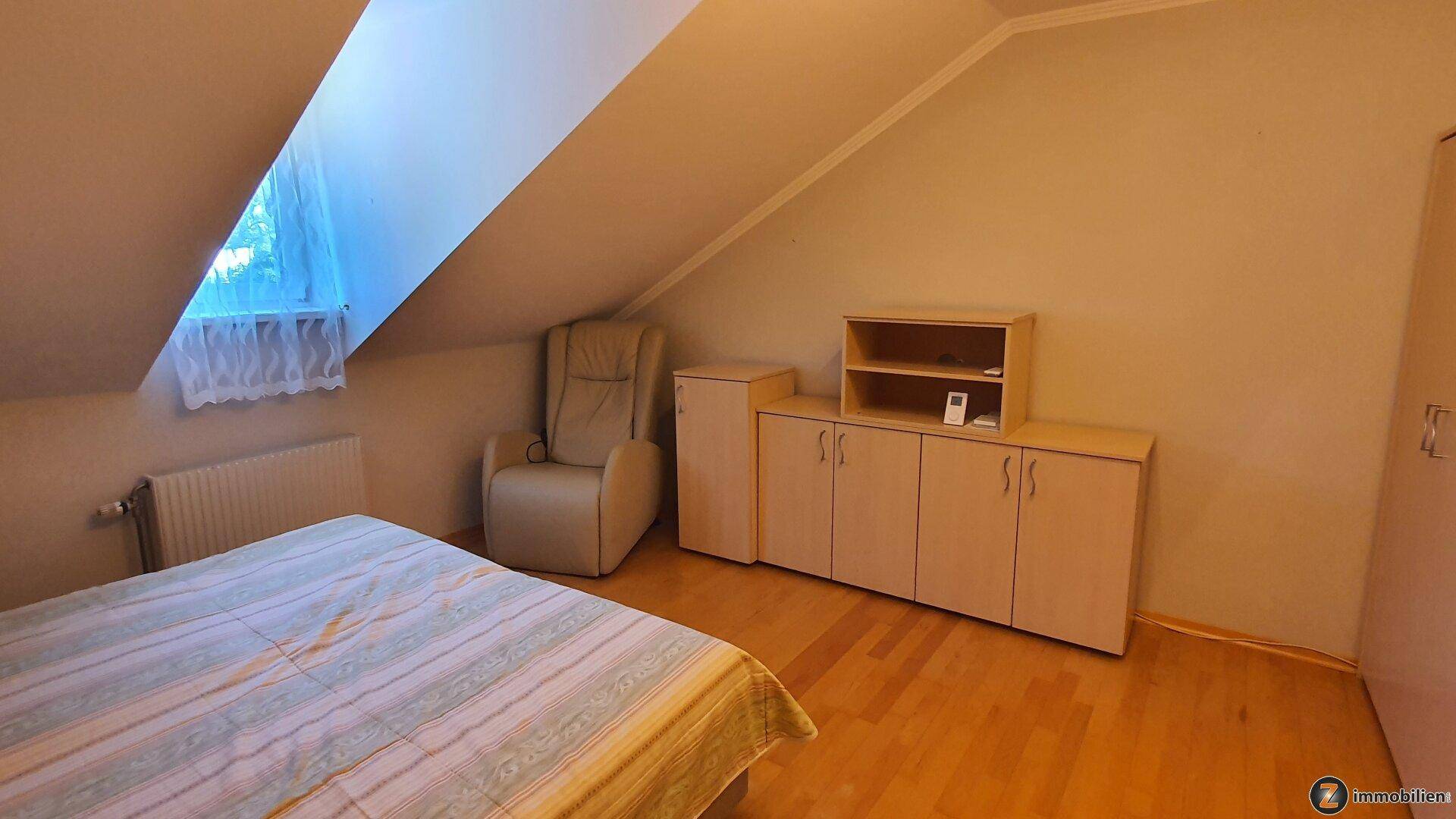 Gemütliches Schlafzimmer im Dachgeschoss mit Holzboden, Schräge und einem bequemen Sessel.