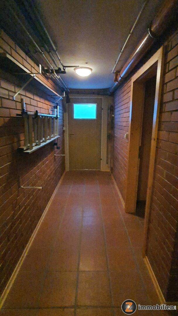 Kellerflur mit unverputzten Ziegelwänden, Fliesenboden und sichtbaren Leitungen an der Decke.