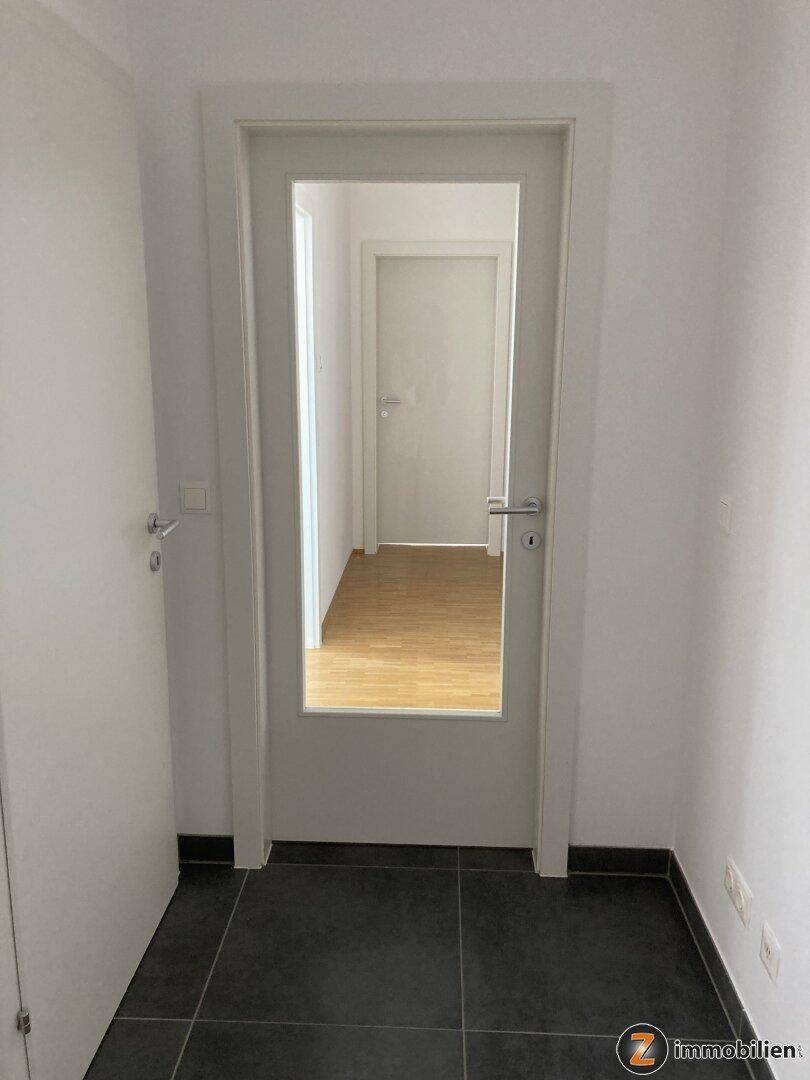 Heller Flur mit dunklen Bodenfliesen und einer Tür mit Glaseinsatz.