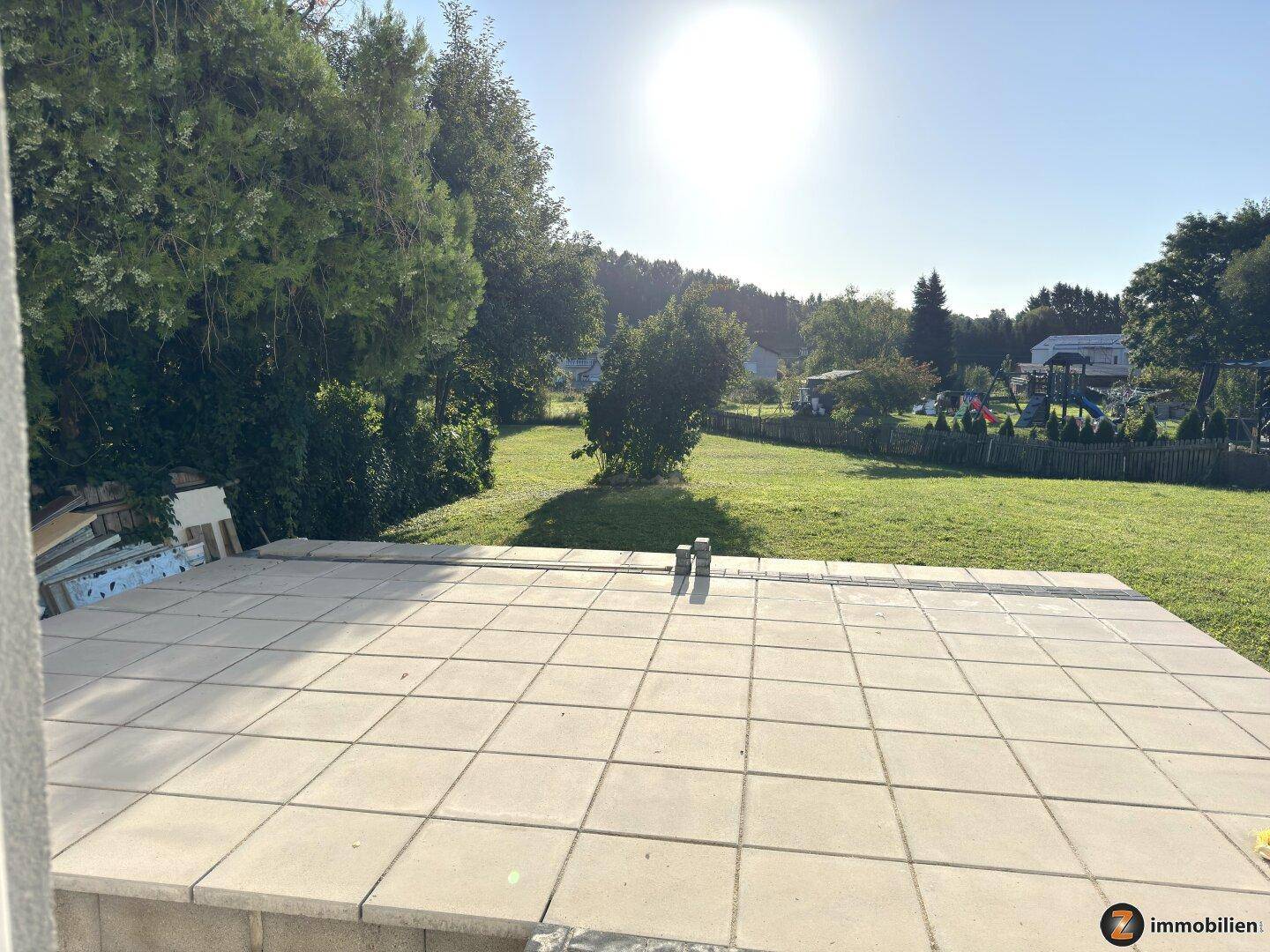 Große gepflasterte Terrasse mit sonnigem Blick auf den weitläufigen Garten und Spielplatz.