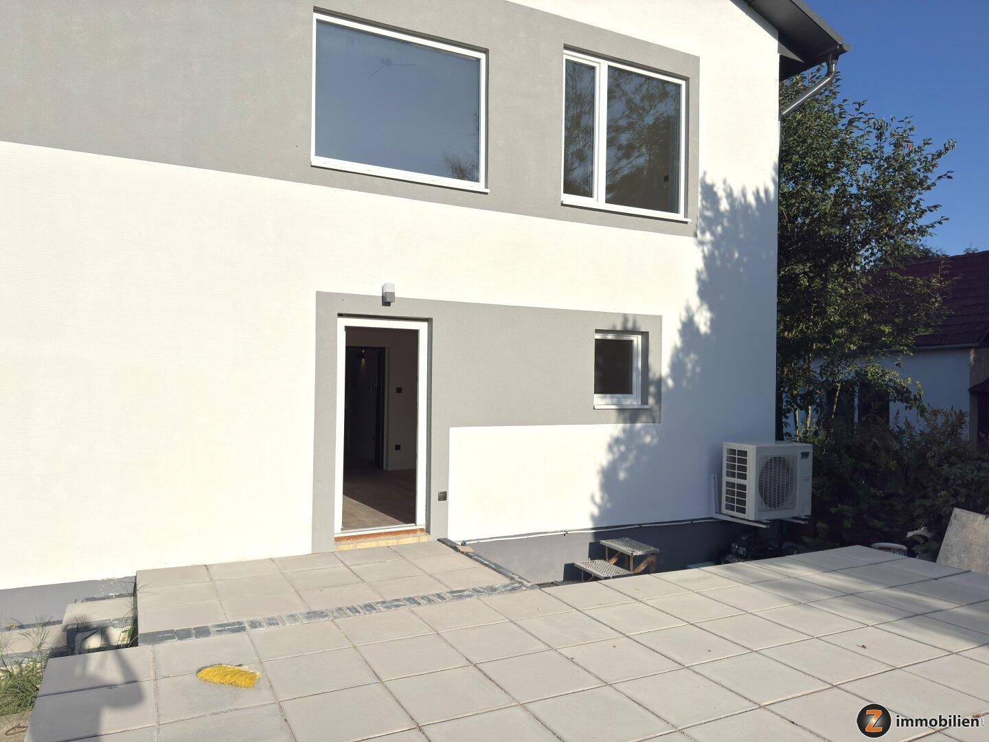Moderne Hausfassade in Weiß und Grau mit gepflastertem Terrassenbereich und Klimaanlage.