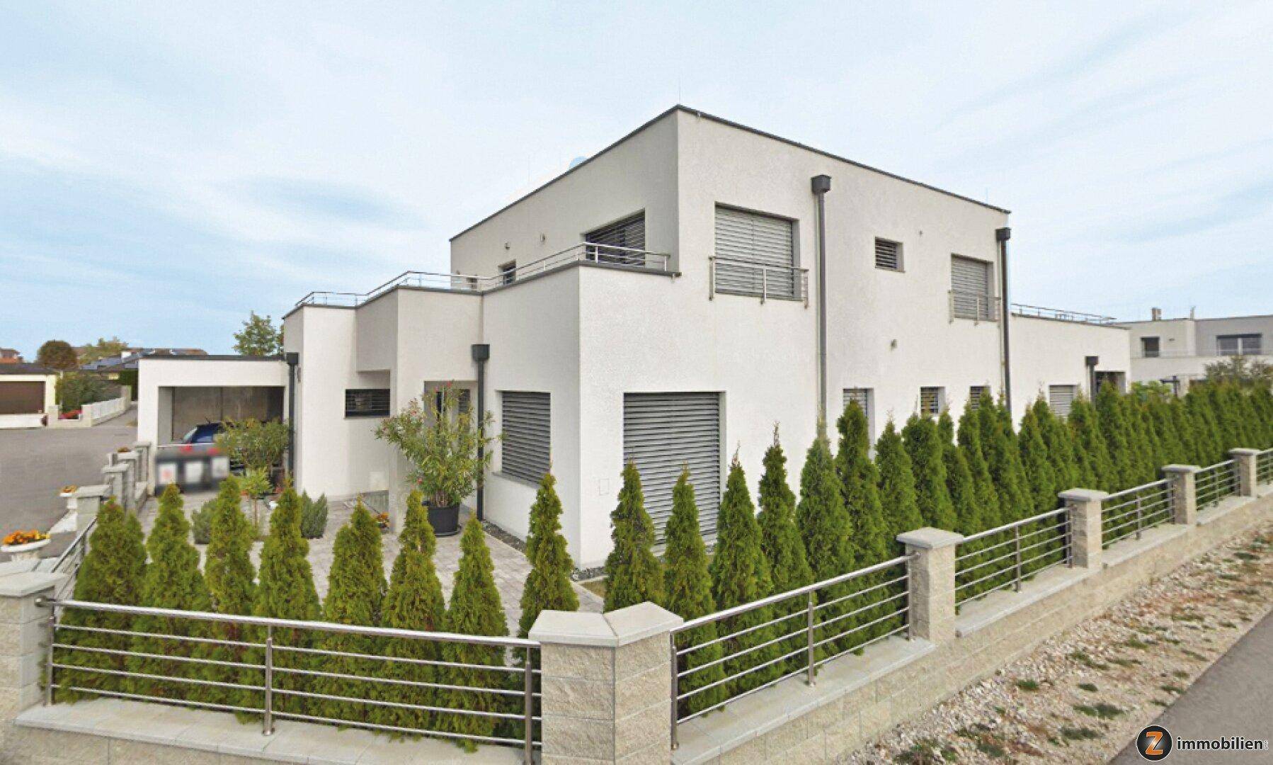 Moderne weiße Villa mit Flachdach, großen Fenstern, Rollläden und gepflegter Hecke im Vorgarten.