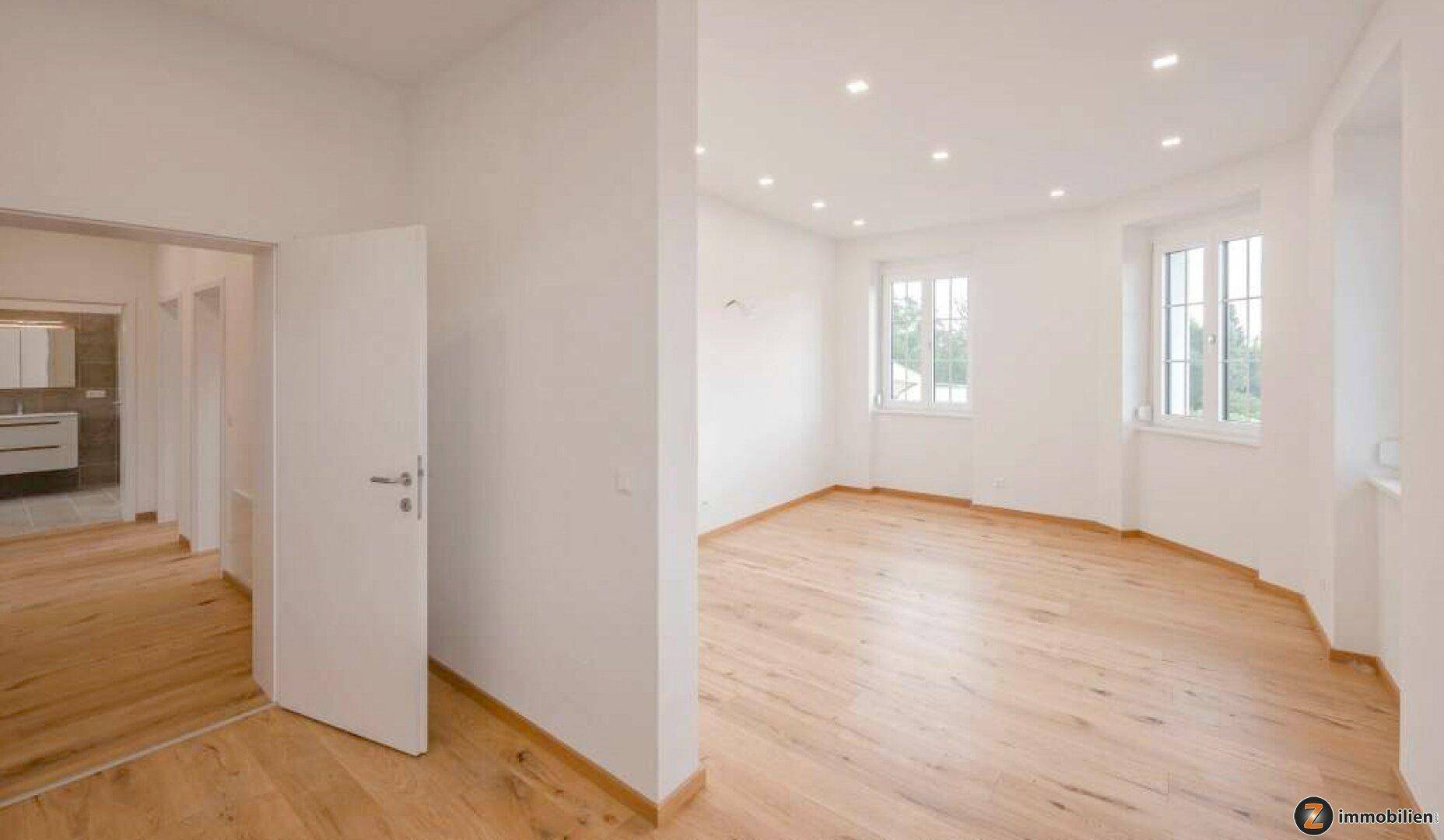 Heller Wohnbereich mit Holzboden, weißen Wänden und Blick in ein modernes Badezimmer.