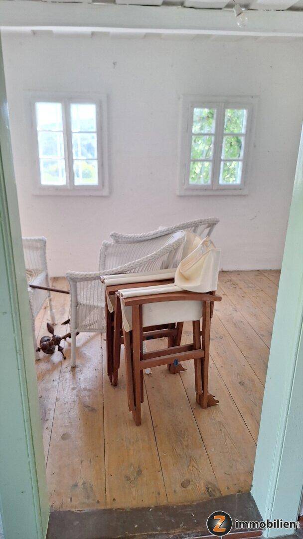 Heller Innenraum mit weißen Wänden, Holzdielenboden und zwei kleinen Fenstern, möbliert mit Korbstühlen.