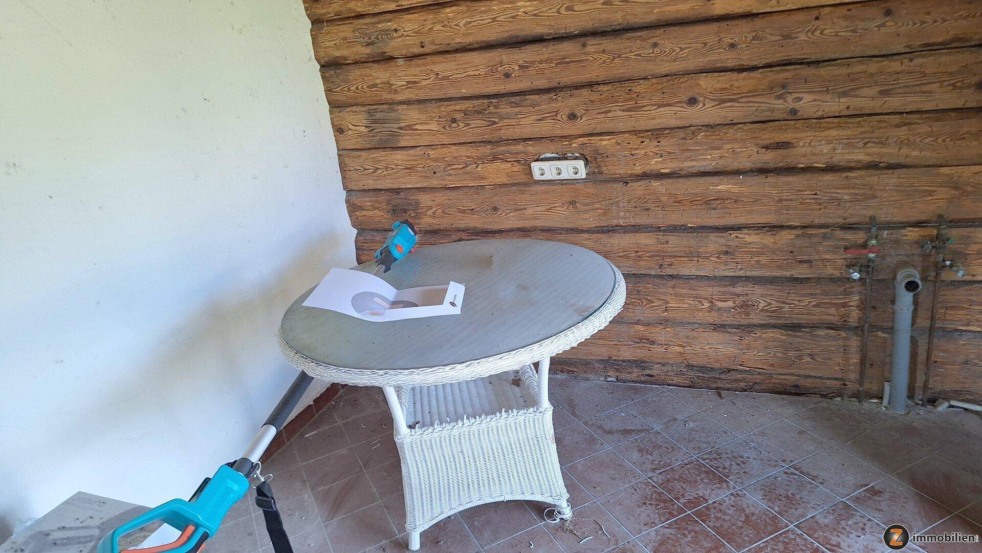 Detailansicht der überdachten Terrasse mit Holzwand, Korbtisch und sichtbaren Installationen.