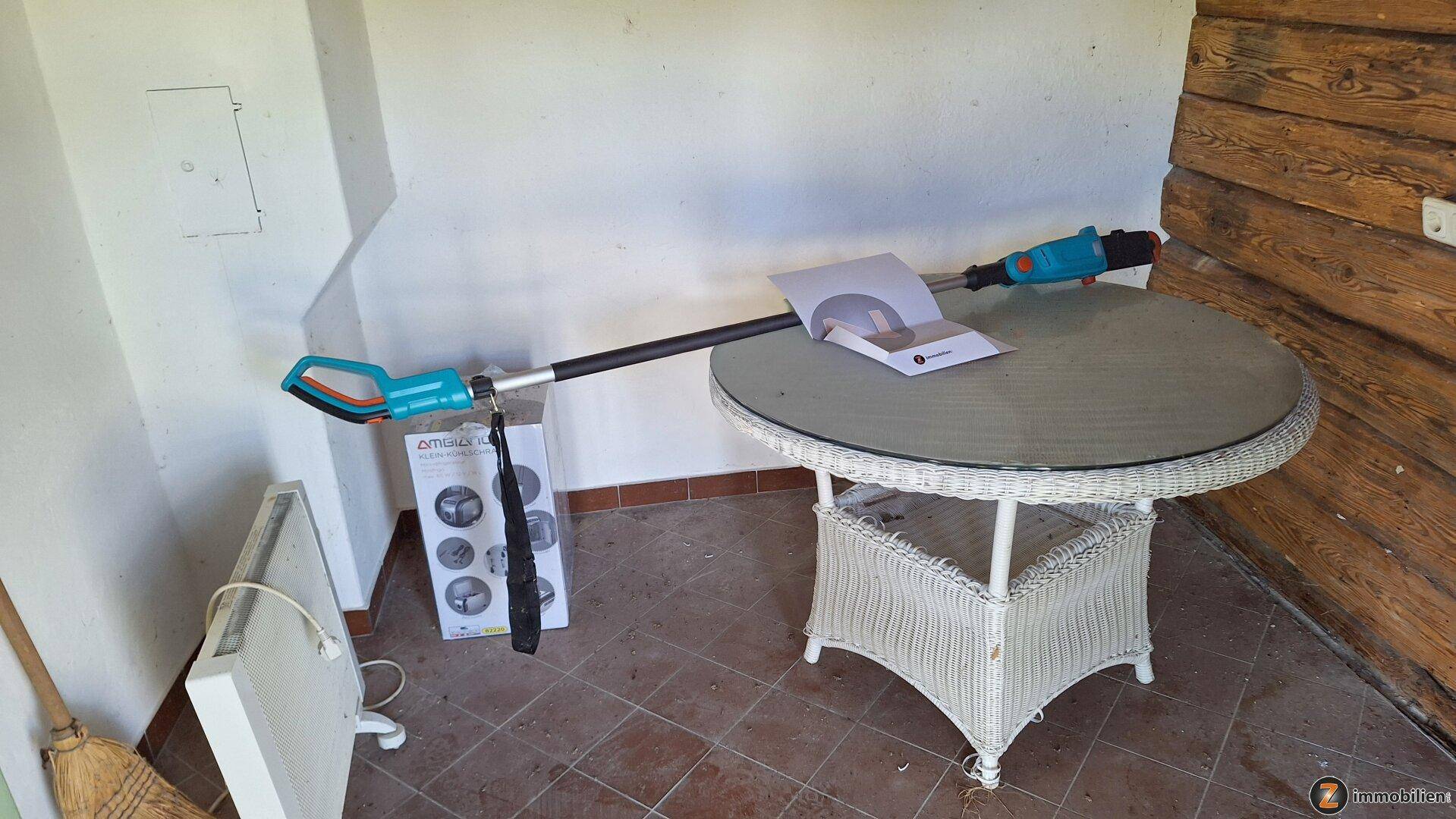 Überdachte Terrasse mit Korbtisch und Gartengeräten, Holzwand und Fliesenboden.