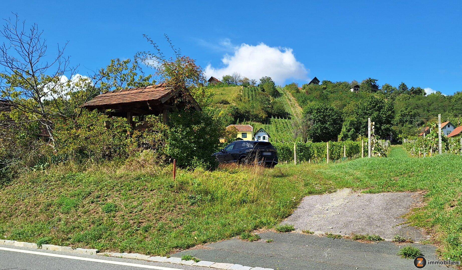Weitläufige grüne Landschaft mit Holzüberdachung, geparktem Auto und Weinbergen am Horizont.