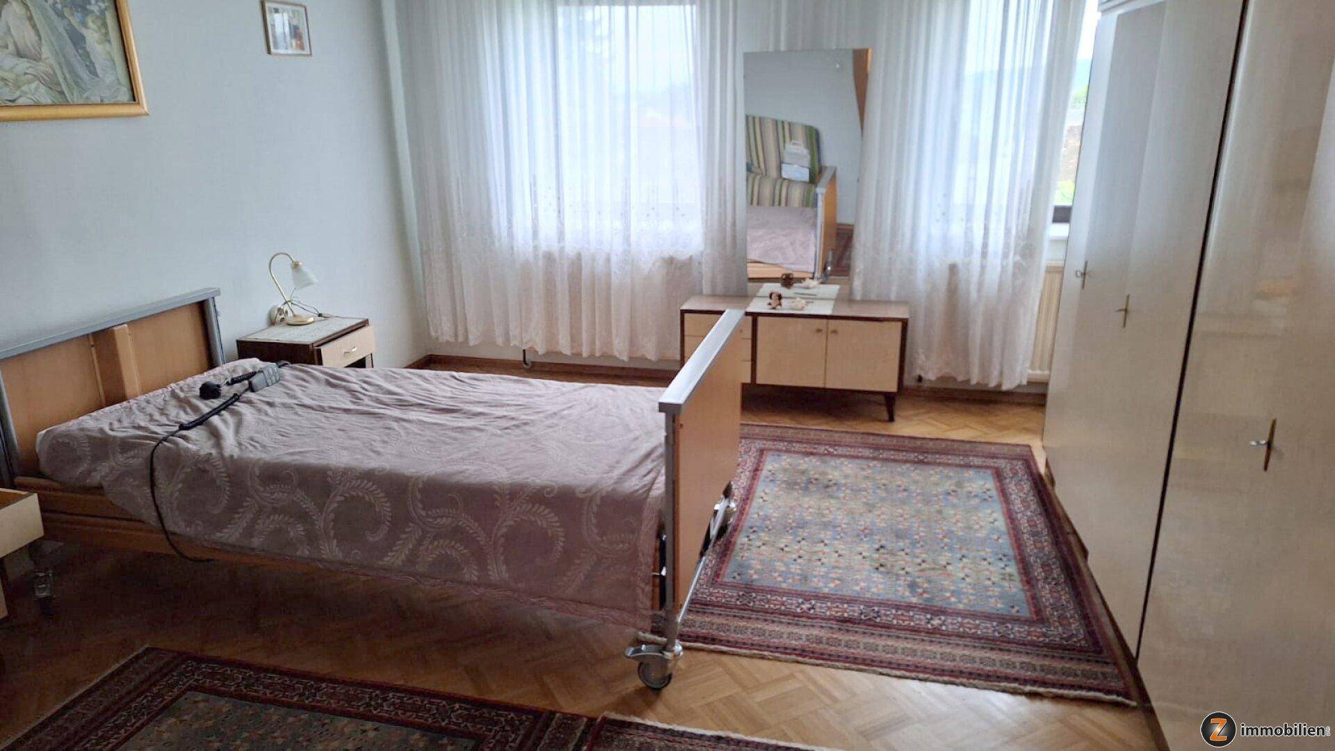 Helles Schlafzimmer mit Einzelbett, Kleiderschrank und großen Fenstern mit Vorhängen.