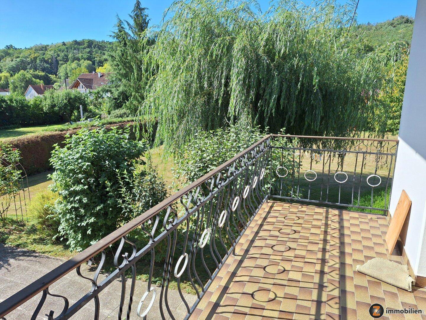 Balkon mit gemusterten Fliesen, schmiedeeisernem Geländer und weitem Blick ins Grüne.