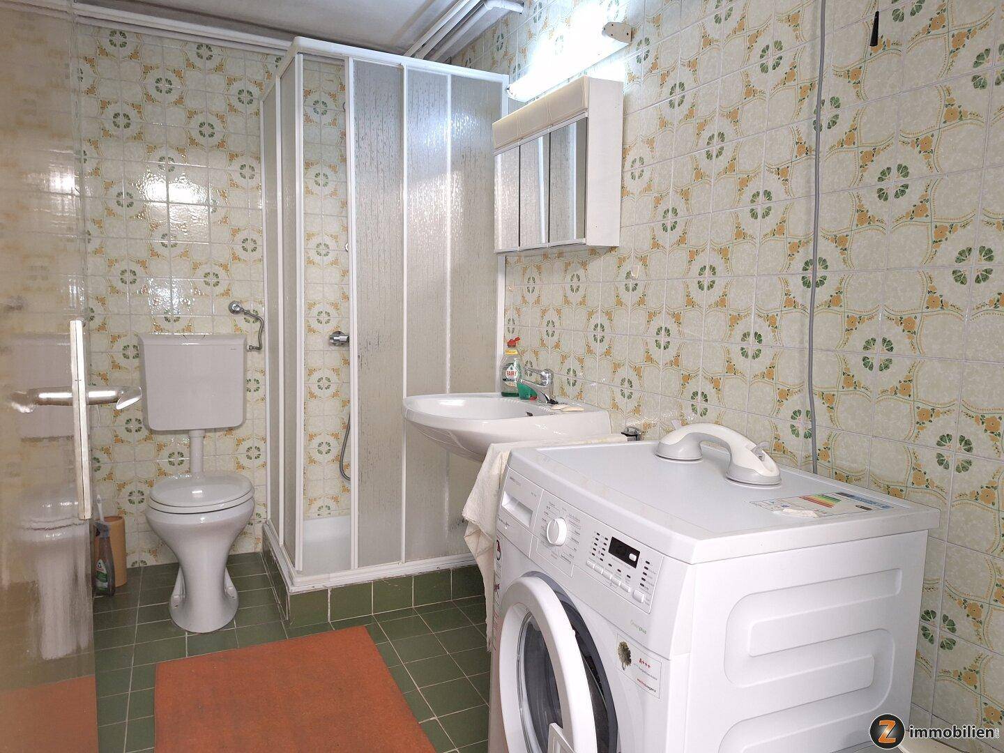 Funktionales Badezimmer mit Dusche, WC, Waschbecken und Waschmaschine, hell gefliest.