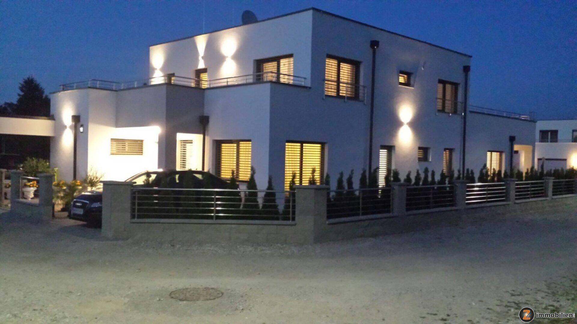 Modernes Einfamilienhaus bei Dämmerung mit beleuchteter Fassade und gepflegtem Vorgarten.