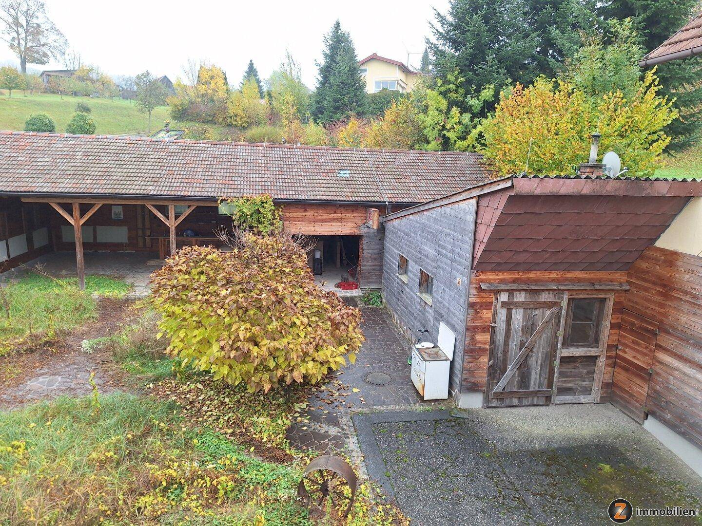 Gartenbereich mit einem rustikalen Holzschuppen und viel Grün, umgeben von Bäumen und Hügeln.