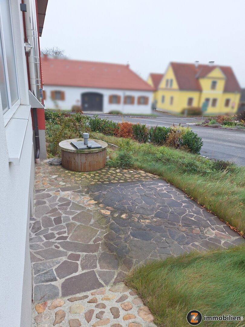Gepflasterter Weg mit einem alten Brunnen und Blick auf weitere Häuser in der Ferne.