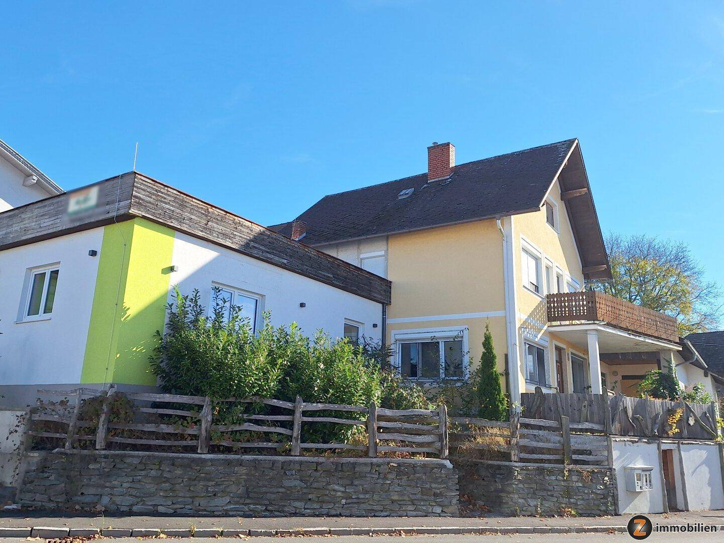 Gelbes Gebäude mit traditionellem Dach und einem Balkon, umgeben von einer Steinmauer und Vegetation.