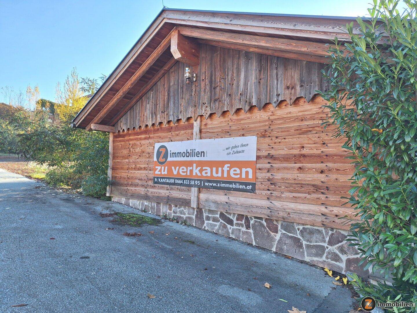 Holzgebäude mit einem 'Zu verkaufen'-Schild an der Fassade, umgeben von Bäumen und Natur.