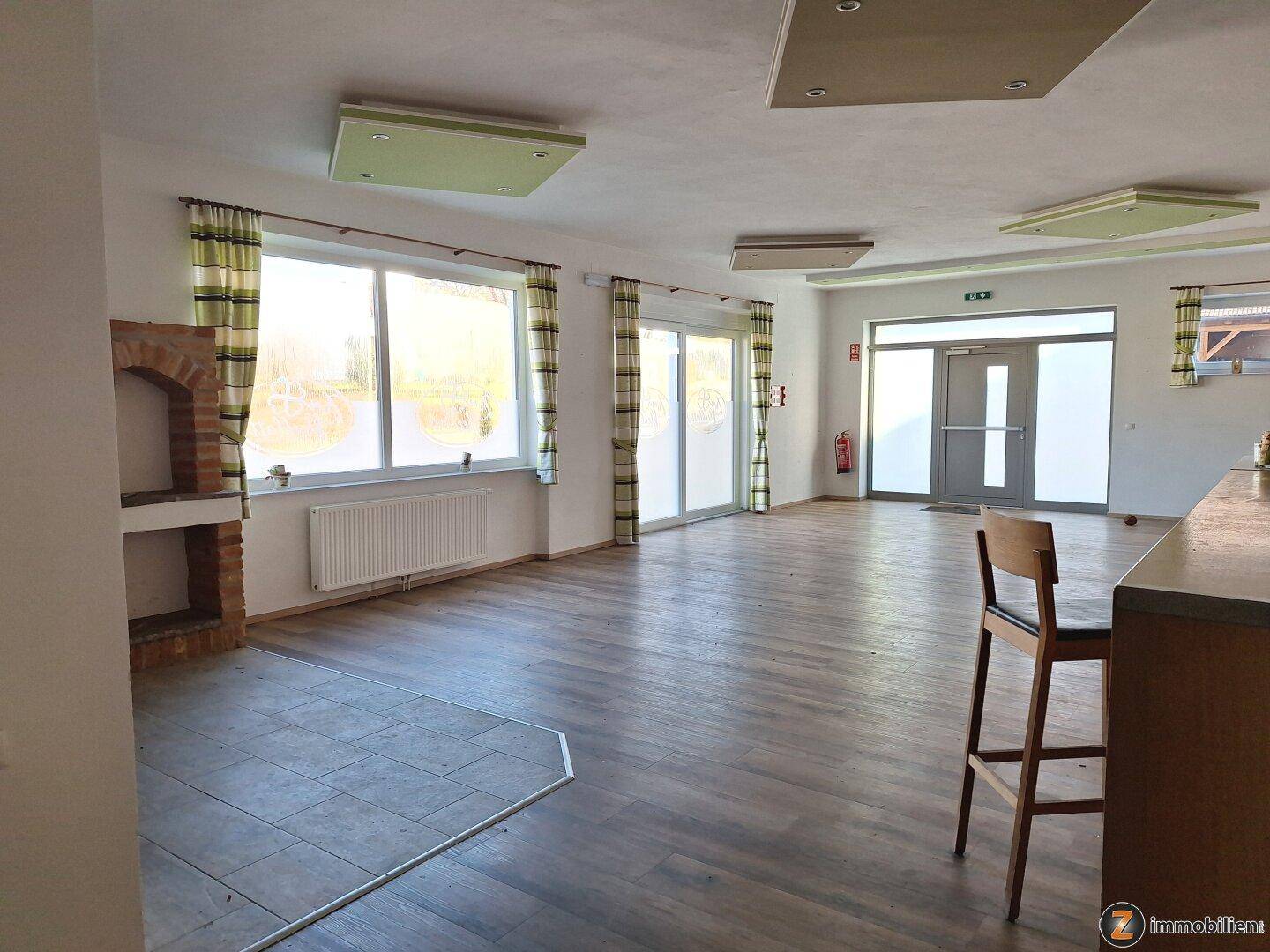 Heller, weitläufiger Raum mit großen Fenstern, Holzboden und einem gemauerten Kamin im Hintergrund.