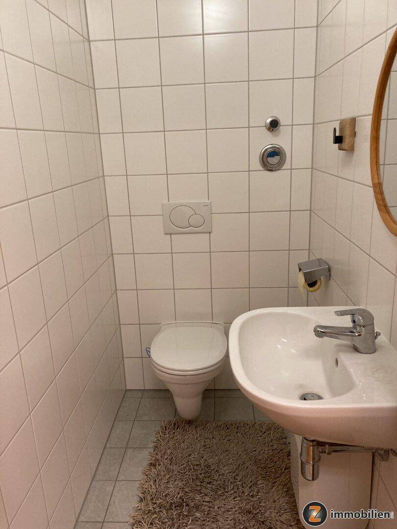 Kleines Badezimmer mit weißen Fliesen, WC, Waschbecken und rundem Spiegel.