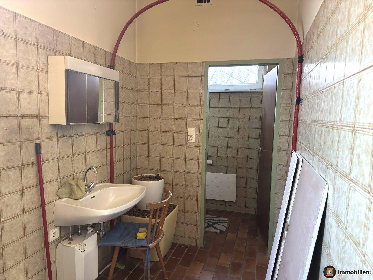 Einfacher Waschraum mit Fliesenwänden und kleinem Waschbecken, angrenzend an eine Toilette.