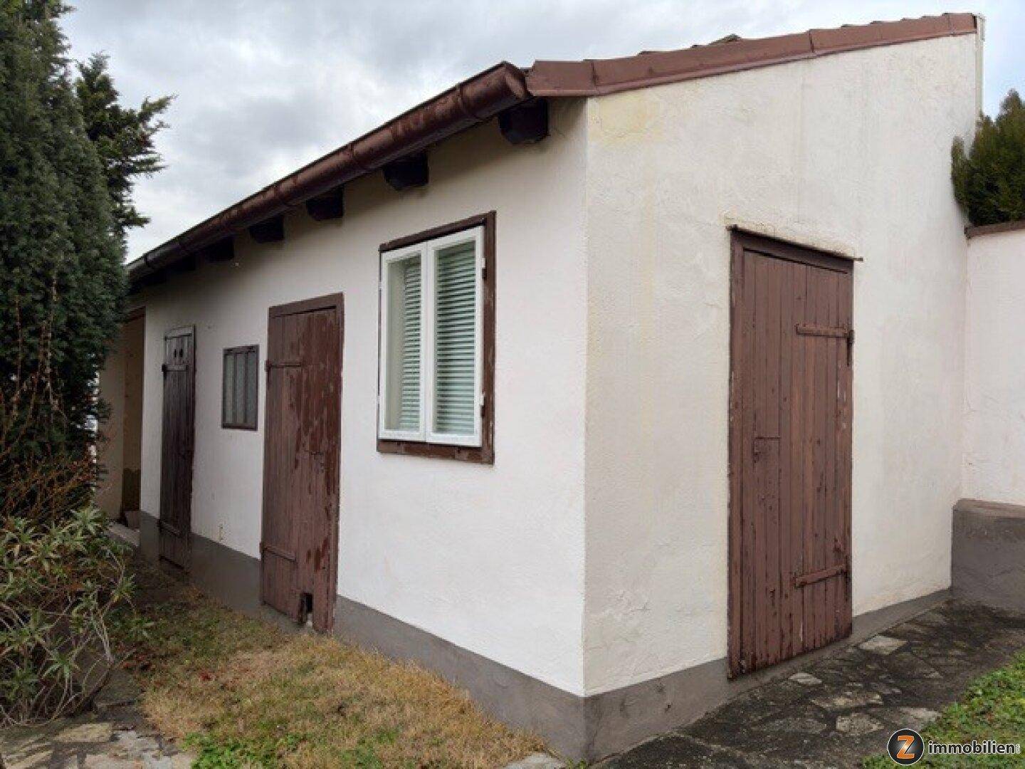 Kleines Nebengebäude im Garten mit weißen Wänden und braunen Holztüren.