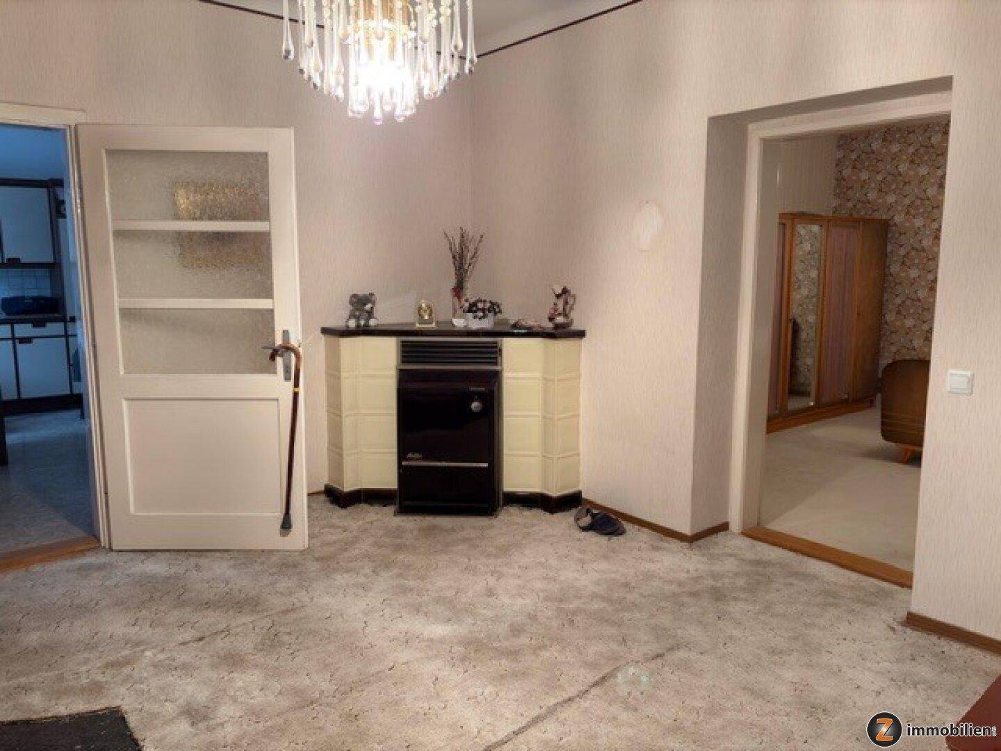 Wohnbereich mit Teppichboden, Kachelofen und Kronleuchter, angrenzend an weitere Räume.