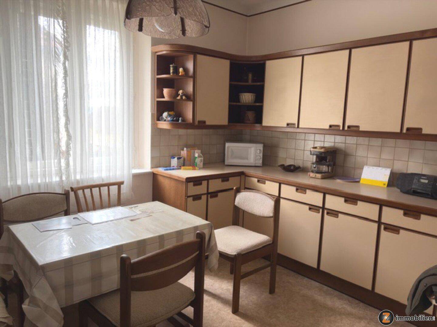 Helle Küche mit beigen Holzschränken, kariertem Tisch und Fenster mit Vorhängen.