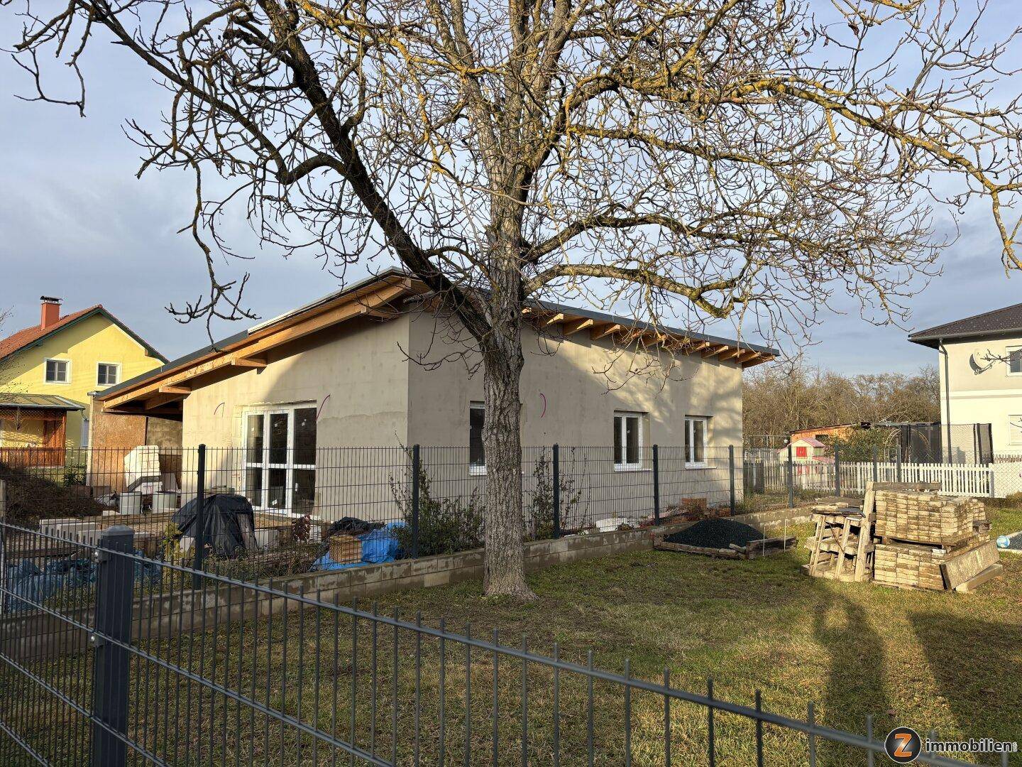 Außenansicht des im Bau befindlichen Hauses mit Gartenbereich, Baum und Baustellenmaterialien im Vordergrund.