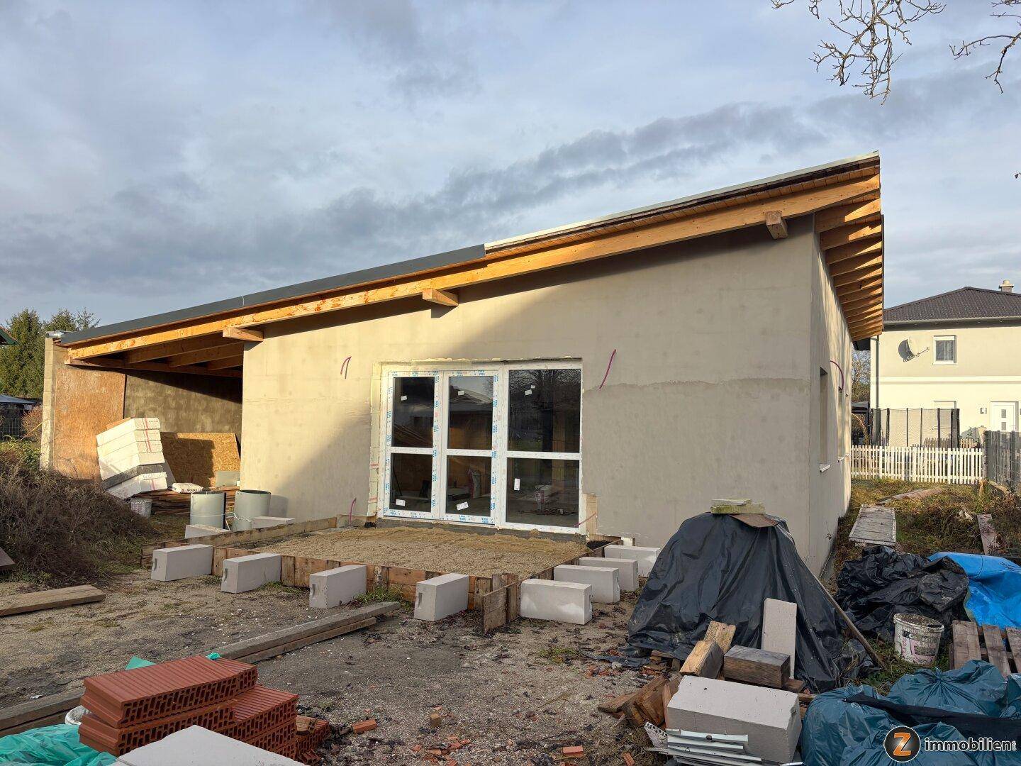 Rückseite des Neubaus mit großer Fensterfront, sichtbarem Dachstuhl und Baustellenmaterialien auf dem Grundstück.