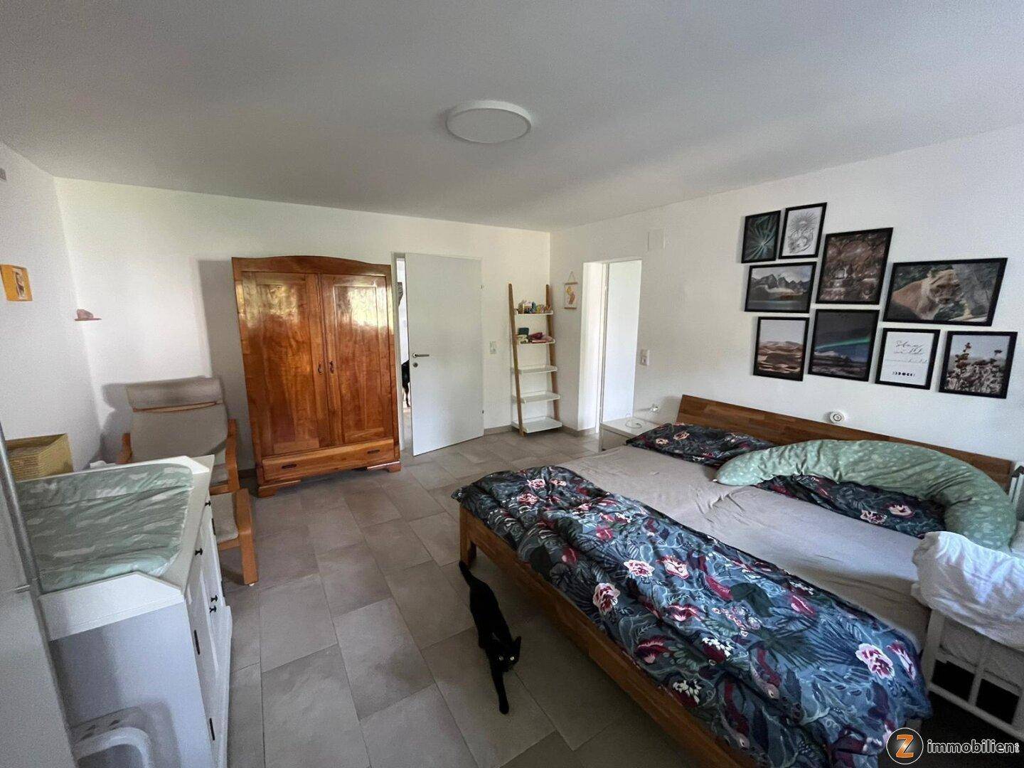 Helles Schlafzimmer mit Fliesenboden, Doppelbett und einem antiken Holzschrank.