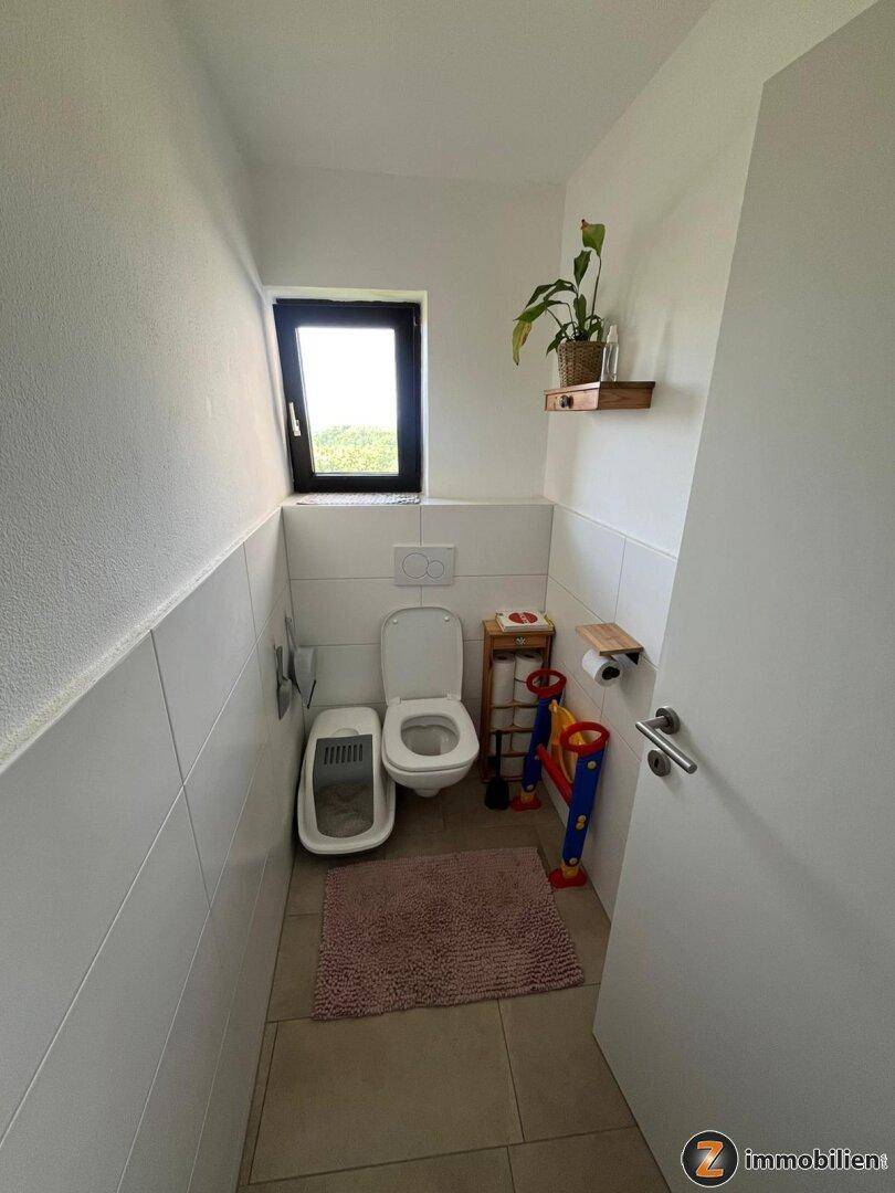 Kleines WC mit Fliesenboden, wandhängender Toilette und einem Fenster für Tageslicht.