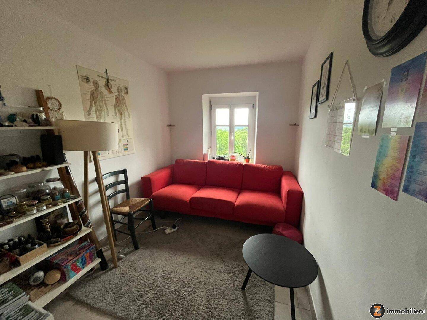 Gemütliches Wohnzimmer mit rotem Sofa, Teppichboden und einem kleinen Fenster mit Blick ins Grüne.