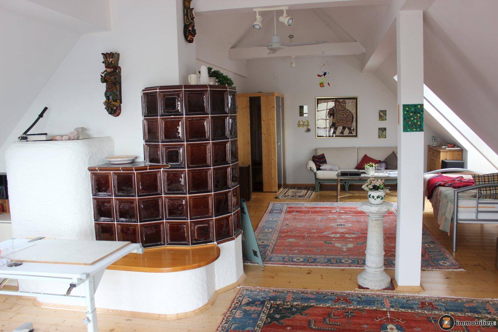 Helles Dachgeschoss-Wohnzimmer mit traditionellem Kachelofen, Holzboden und gemütlichen Teppichen.