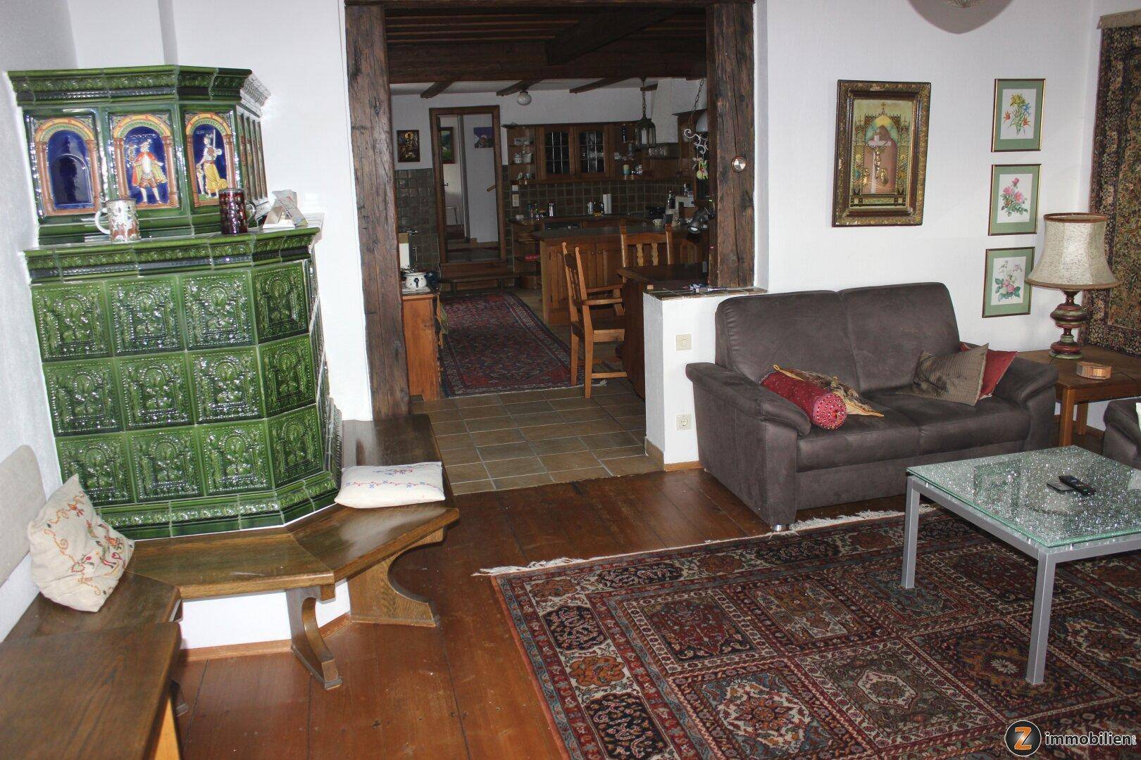 Gemütliches Wohnzimmer mit grünem Kachelofen, Holzbalken und Blick in den Essbereich.