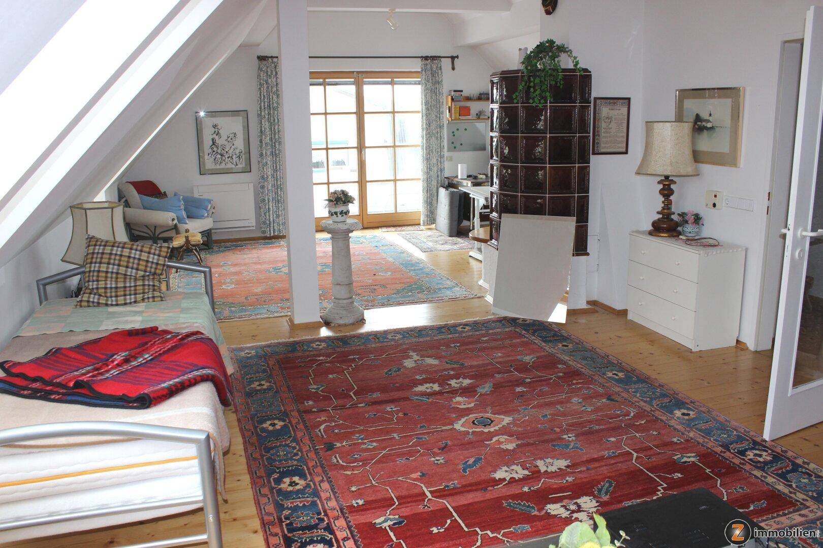 Helles Dachgeschoss-Wohnzimmer mit Kachelofen, Holzboden und Zugang zu einem Balkon.