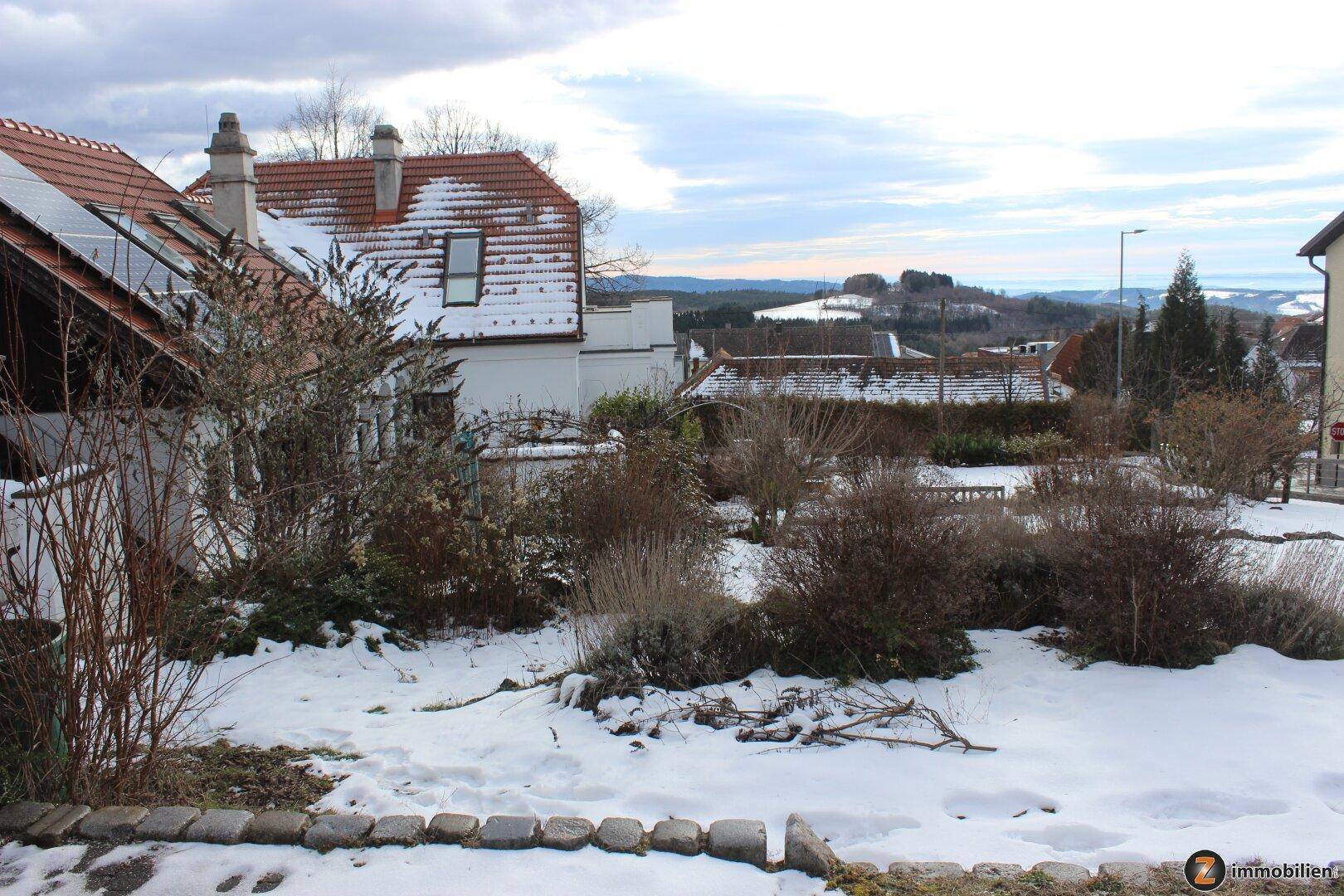 Verschneiter Gartenbereich mit Sträuchern und Blick auf die umliegende Landschaft.