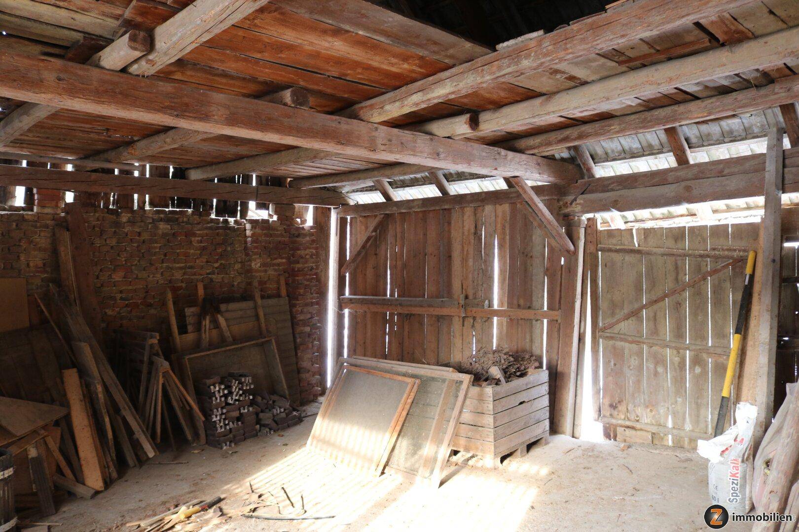 Rustikaler Innenraum einer Scheune mit Holzbalken, Ziegelwänden und gelagertem Holz.