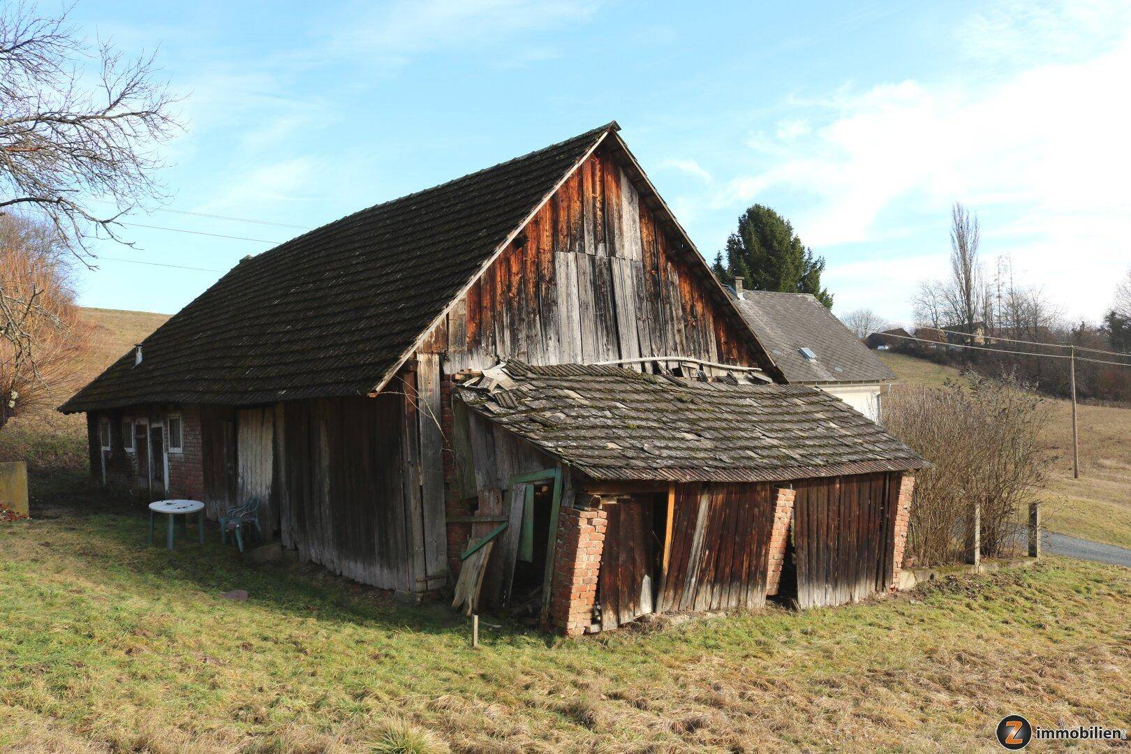Alte Holzscheune mit verwitterter Fassade und baufälligem Anbau in ländlicher Umgebung.