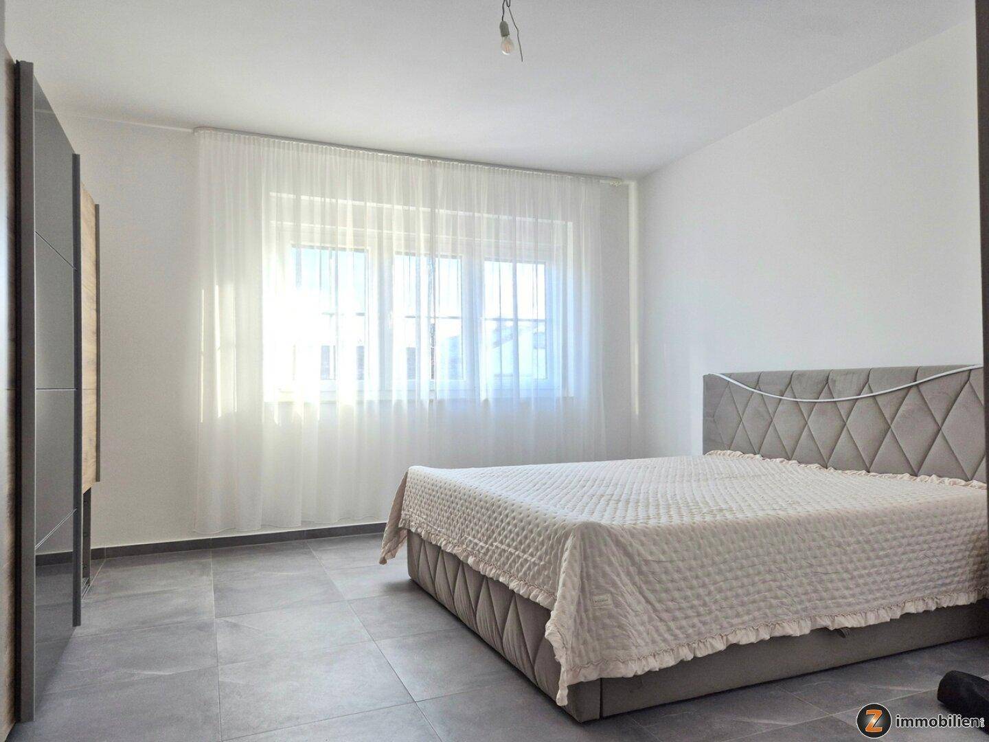 Schlafzimmer mit großem Bett, Fliesenboden und hellen Vorhängen vor dem Fenster.