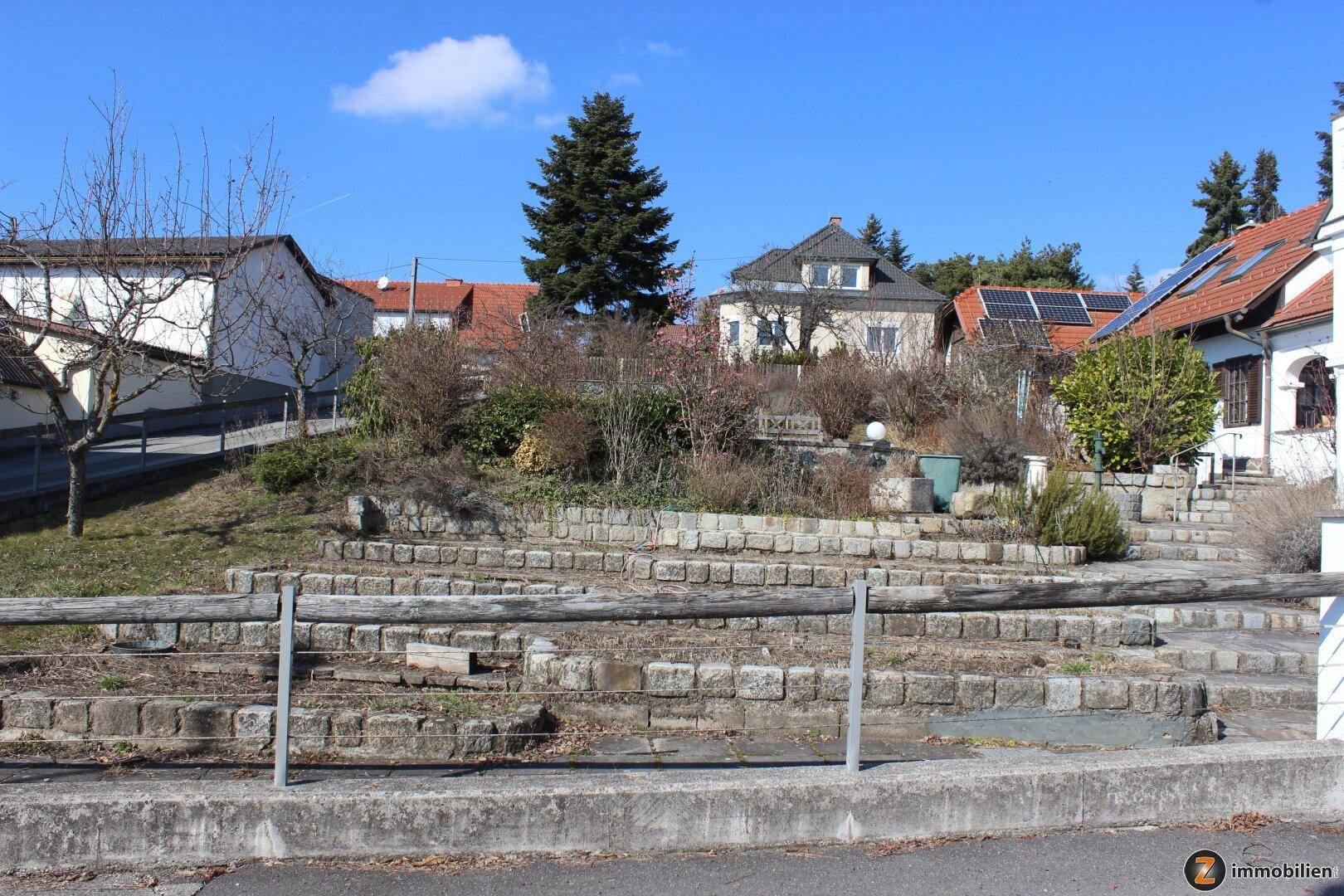 Terrassierter Gartenbereich mit Steinmauern und Holzgeländer, von der Straße aus sichtbar.