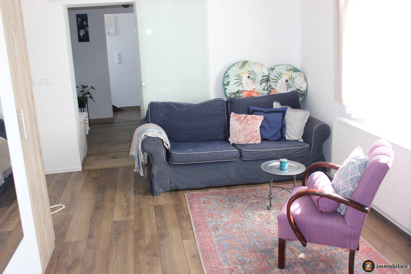 Komfortabler Wohnbereich mit blauer Couch, lila Sessel und Holzboden, verbunden mit einem Flur.